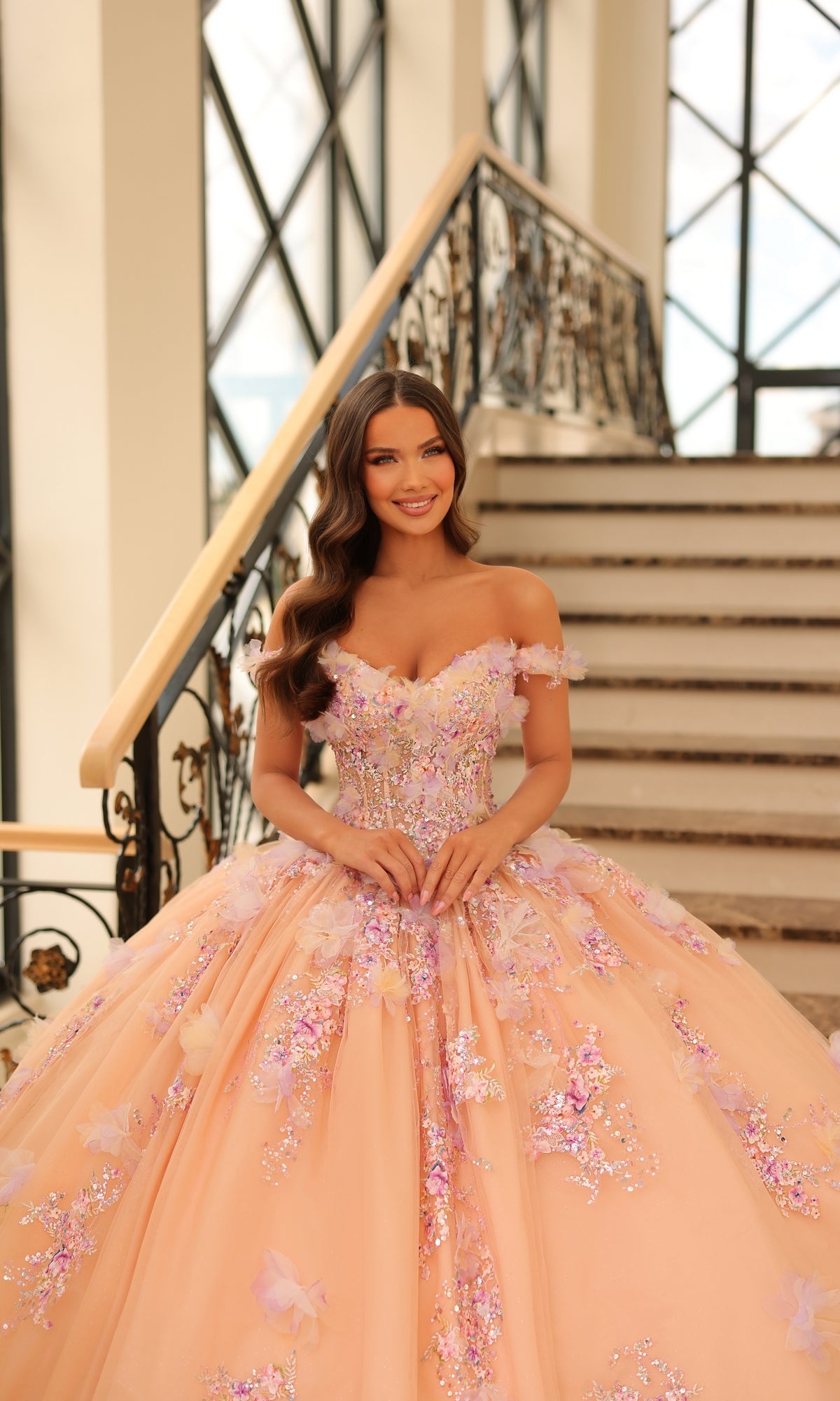 A woman smiles at the camera in front of a staircase, wearing the Quinceanera Dress 54228 By Amarra—an off-the-shoulder, peach ball gown adorned with 3D floral appliqué.